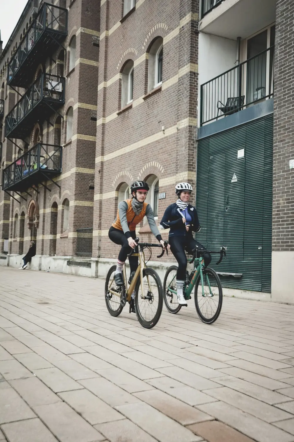 Two cyclists riding side by side past a brick warehouse